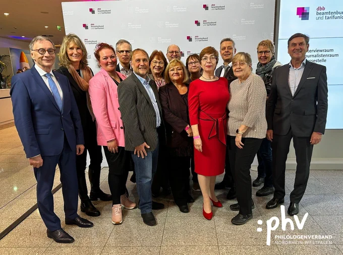 : Das Bild zeigt eine Gruppe von 14 Personen, die sich zu einem Gruppenfoto versammelt haben. Im Hintergrund ist eine Wand mit dem Logo des „dbb beamtenbund und tarifunion“ zu sehen. Die Personen stehen in zwei Reihen: eine vordere und eine hintere. Alle lächeln in die Kamera. In der vorderen Reihe tragen einige Personen auffällige Kleidung, darunter eine Frau in einem roten Kleid. Die anderen tragen formelle oder legere Kleidung, darunter Anzüge, Blazer und Pullover. Das Logo des „Philologenverband Nordrhein-Westfalen“ ist in der unteren rechten Ecke eingeblendet. Die Atmosphäre wirkt professionell und herzlich.