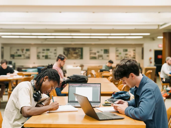 Two students studying in a library, one writing in a notebook and the other using a laptop, with others in the background.