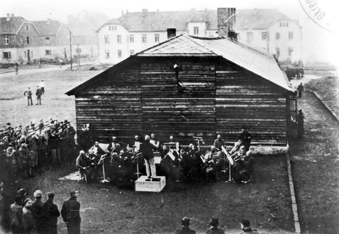A historical photograph showing a group of musicians - camp orchestra at Auschwitz - playing in front of a wooden building, with an audience of individuals standing and observing.
