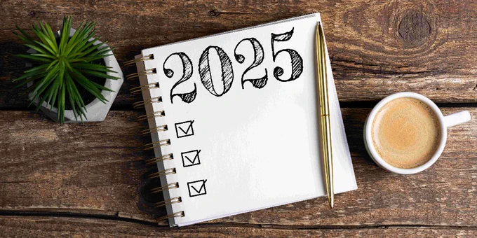 A white notepad on a wooden table with 2025 written on it, and boxes below for a to do list, with ticks in them. A coffee cup and plant are on either side of the notebook.