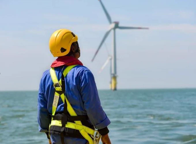 A person in a hard hat and harness looking at a wind turbine in the distance.