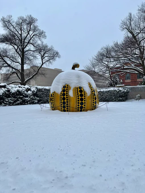A Yayoi Kusama pumpkin sculpture adorned with black polka dots. The pumpkin is yellow and huge. It is displayed outdoors on a snow-covered ground.