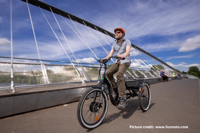 Man wearing a shirt and trousers and a helmet and sunglasses is cycling a black trike across a walking, wheeling and cycling bridge