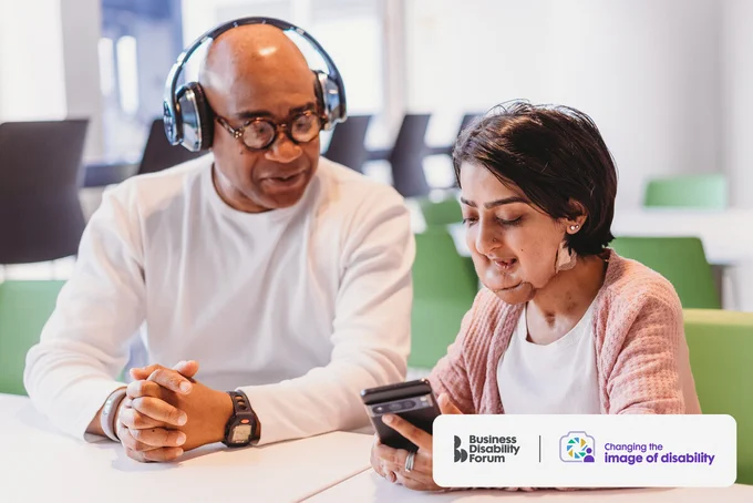 Person with a facial difference who is short in stature sits looking at their phone next to a colleague who is wearing headphones. The Business Disability Forum 'Changing the image of disability' logo is in the lower right corner of the image.