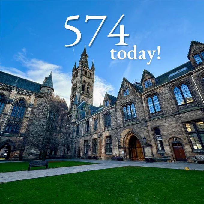 The Gilbert Scott building at the University of Glasgow, featuring its iconic tower and spire and gothic-style architecture under a clear blue sky, with green lawns in the foreground. Text overlay reads '574 today!'