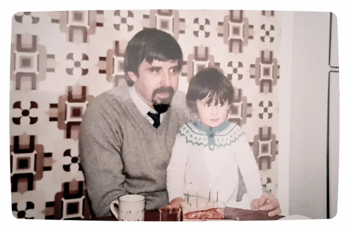 A photo of me and my dad at a kitchen table in front of some glorious brown and 70s wallpaper. He has dark hair and a dark goatee, and is wearing a grey v-neck sweater and a shirt and tie. I'm around 3 or 4 years old with a mop of dark hair and a white winter sweater, sitting on his knee and looking a bit worried.