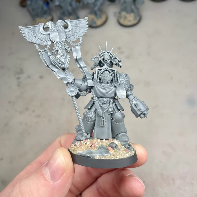Custom Grey Knight Terminator/Paladin Ancient conversion, unpainted and crafted by Andy TheBrushSmith. This work-in-progress model features a detailed banner with a winged eagle motif, paired with iconic Grey Knight armor elements, purity seals, and decorative chains. The model is further enhanced by a striking skull-adorned halo and intricate details on its armor. Its scenic base is designed with realistic rubble, textured earth, and debris, adding to the dynamic battlefield aesthetic. Perfect for Warhammer 40K enthusiasts seeking unique and inspiring miniature conversions.
