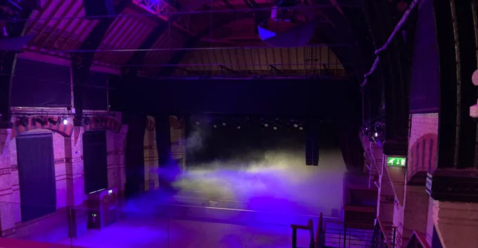 The image shows the interior of the Cambridge Corn Exchange with a seemingly empty stage and venue.