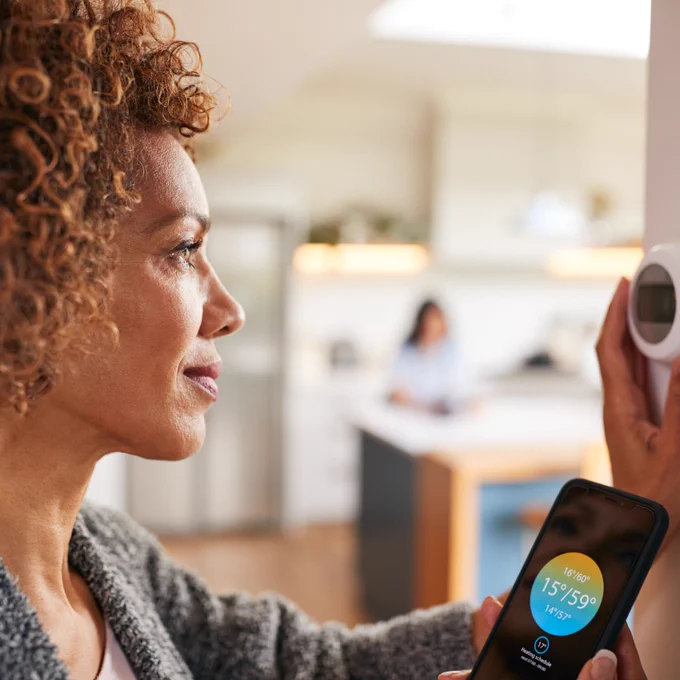 A woman with curly brown hair looks at her smart thermostat feature on her phone and adjusts the appliance on the wall.