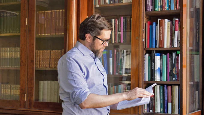 An IASH Fellow in the Fellows' Library: a man with dark hair, glasses and a beard wearing a blue shirt. He is reading a book in front of glass-fronted bookshelves.