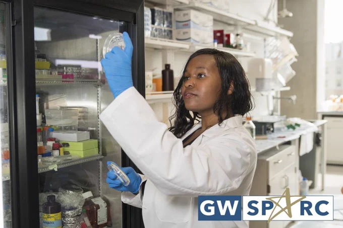 A researcher in a white lab coat examines a petri dish in a laboratory with GW SPARC logo on bottom right.