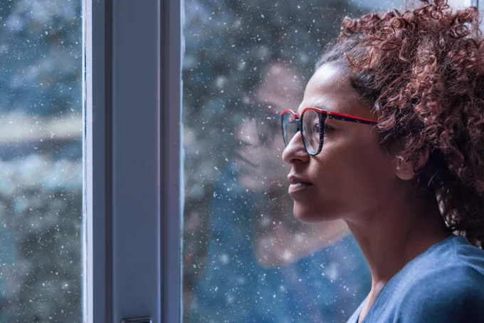 Lonely African American woman staring out a window with a sullen face as snow falls outside