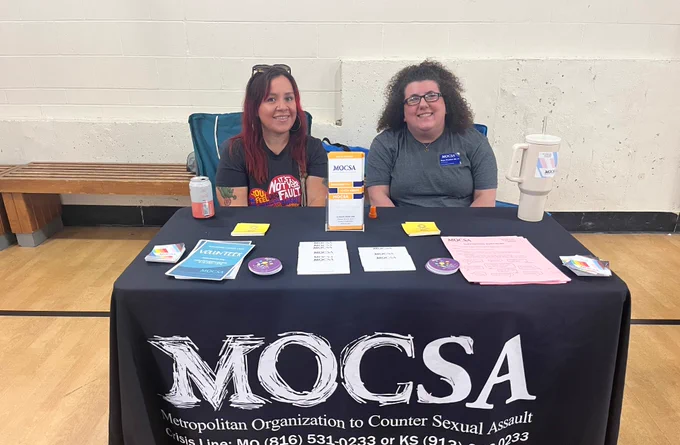 Two MOCSA staff members sit behind a table with a black table cloth with white text that reads MOCSA. 