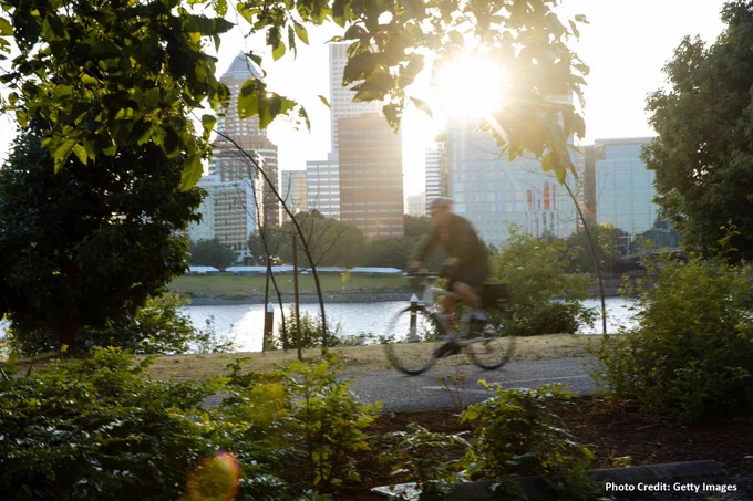 Person cycling on a path by a river at sunset with city skyline in the background. Photo credit: Getty Images.

