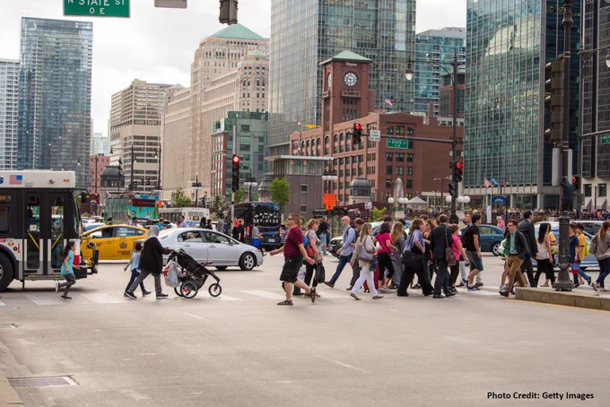Pedestrians crossing a busy intersection in downtown Chicago with skyscrapers and traffic in the background. Photo credit: Getty Images.
