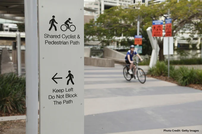 A sign indicating "Shared Cyclist and Pedestrian Path" with directional arrows and instructions to "Keep Left Do Not Block The Path." In the background, a person on a bicycle is riding along the designated path.
