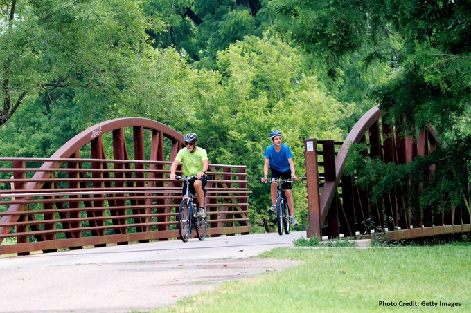 Two cyclists riding bikes over a wooden arch bridge in a lush park.
