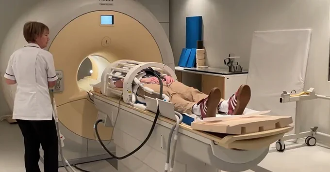 A healthcare professional stands beside an MRI machine, monitoring a person who is lying down inside the scanner for a medical examination.