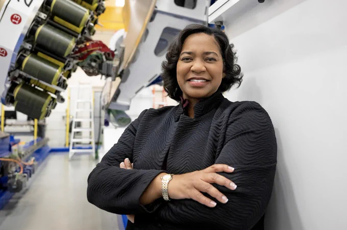 Tawnya Laughinghouse is standing in a high-tech industrial facility, smiling and crossing her arms. She is wearing a textured black jacket and a gold bracelet. Behind her are large pieces of machinery and industrial equipment, with bright lighting highlighting the space. Credit: NASA/Charles Beason
