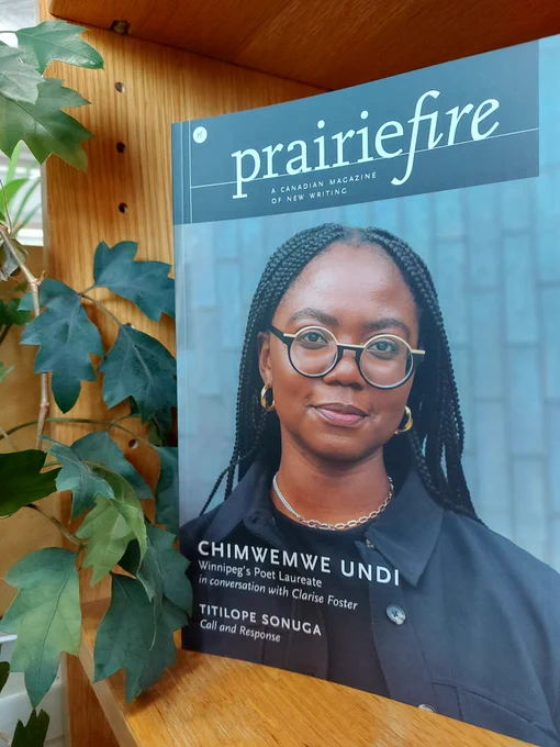 Photo of a bookshelf with the latest issue on it next to a Kangaroo fern. Issue is a dark, charcoal blue, with a photo of Chimwemwe, with braided hair, round glasses that are light yellow on top and black at the bottom, gold earrings, a silver necklace and a dark blue shirt and jacket. She is slightly smiling. 