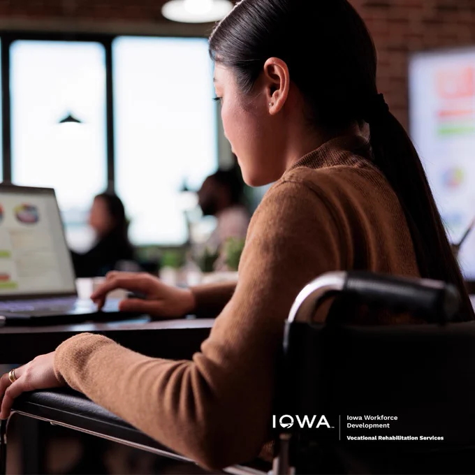 A woman sitting down at a desk using a computer. The image also features the Iowa Vocational Rehabilitation logo. The woman is also sitting in a wheelchair in the image. 