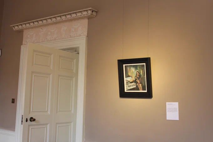 The oil painting 'Estranged Lady Cooking Stew' by Juliette Morrison hangs on the wall to the right of a large white door that is ajar. The painting depicts a woman sitting cross-legged beside a stove that has an active fire in a pot. She seems contemplative and indifferent to the fire roaring beside her. The background features muddy, abstract colours, creating an unsettling atmosphere.