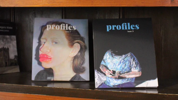 The image shows two literary journals on a wooden shelf, Profiles (issue 2) and Profiles (issue 3). The cover on the left features a woman with enlarged lips and a piercing stare. The one on the right depicts a figure seated on the edge of a bed and holding a device, but their head is obscured by the darkness.