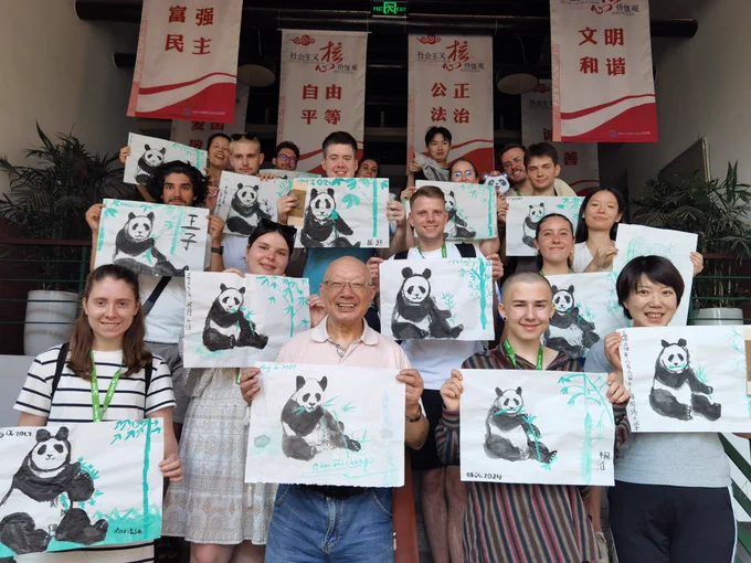 A group of people holding paintings of pandas, standing on a staircase. The artwork features black and white pandas with green bamboo, created using traditional brushstroke techniques. Behind them, banners in Chinese characters are displayed. The participants of the cultural workshop appear diverse, smiling and proud of their creations.