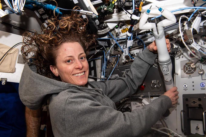 iss070e097708 (Feb. 21, 2024) --- O’Hara is wearing a gray hooded sweatshirt and smiling at the camera, as she holds the miniature robot with both hands. The robot is narrow, about the size of an adult’s forearm, with a gray handle that has up and down arrow buttons and two bent “arms” on its end. One of the arms has a metal spatula and the other a set of clippers.