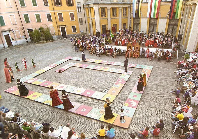 People in medieval dress standing on a giant game board in a town square. 