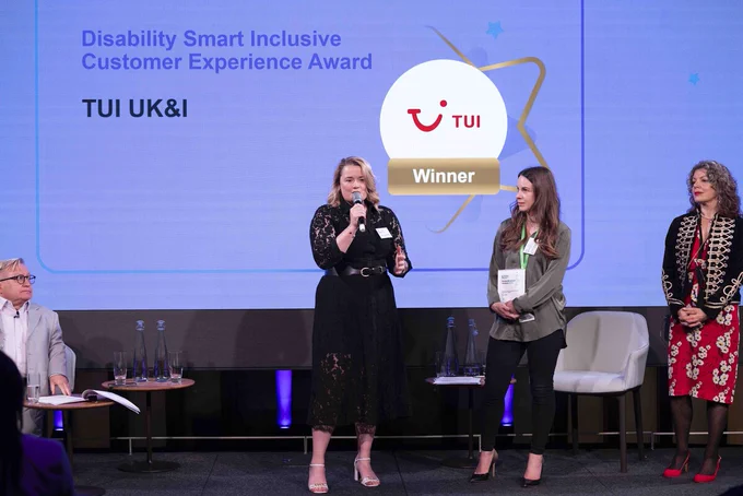 4 people on stage at an event. The person in the centre is speaking into a microphone, and the person to their right is holding an award. Behind the group is a screen with the words 'Disability Smart Inclusive Customer Experience Award'.