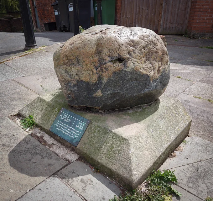 A large boulder on a plinth, with a plaque on the front. There is a fence and some wheelie bins in the background. Photo from August 2024.