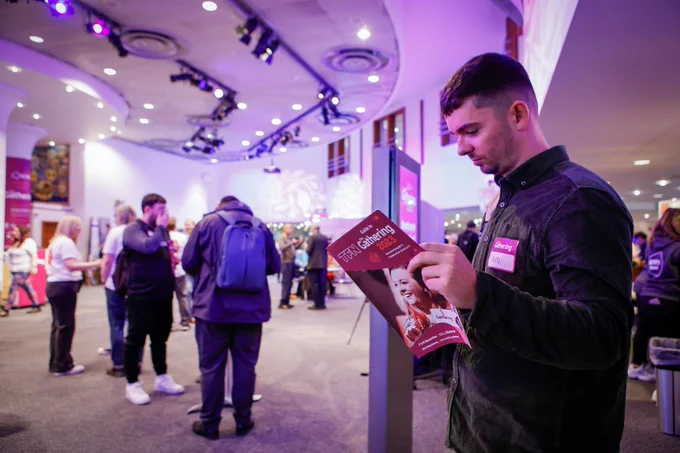 A delegate wearing a dark shirt at the Gathering stands side-on reading the Gathering guide at the EICC in Edinburgh. In the background out of focus are several people standing in small groups speaking to one another. 