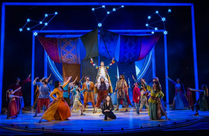 Performance of 'Joseph and the Amazing Technicolor Dreamcoat'. In the centre, a man (Adam  Filipe as Joseph) is standing on someone else's shoulders and is wearing a long rainbow coloured coat. Around them stand a large group of cast all wearing brightly coloured costumes and pointing at Joseph.