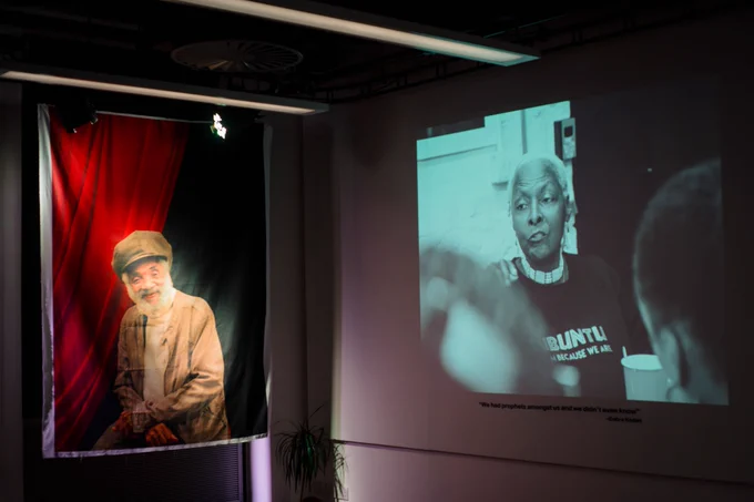 Two walls in a dimly lit exhibition space. The left wall has a large piece of hanging cloth with the image of a bearded man sat against a black and red background. The right wall has a black and white projection of a person seemingly mid-speech.