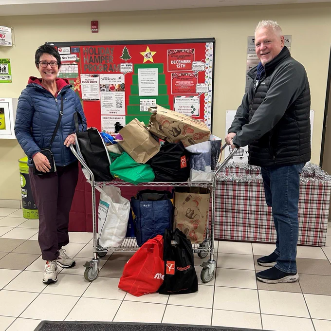 Photo of 2 adults standing with a cart filled with bags of non-perishable food donations.