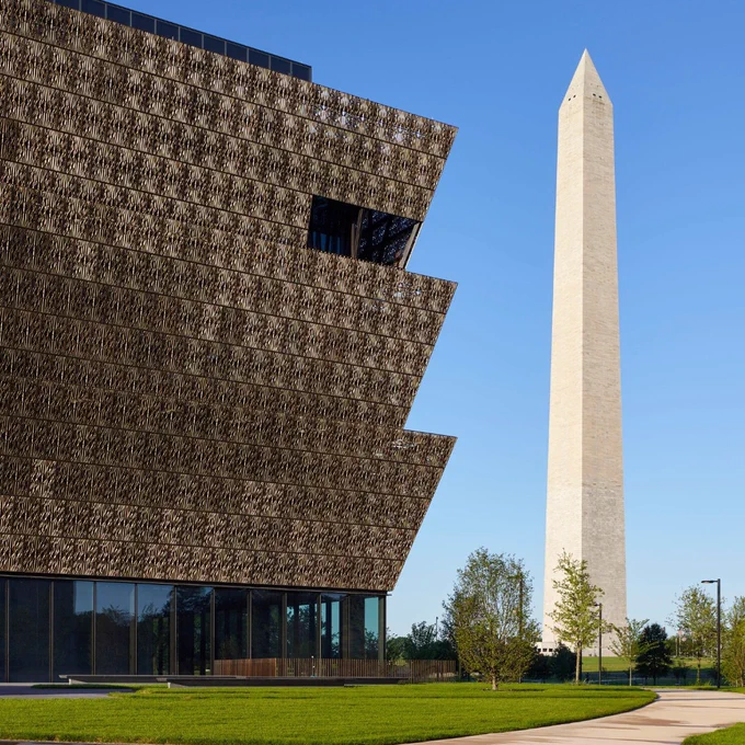 Exterior view of the National Museum of African American History and Culture with its distinctive tiered design, next to the Washington Monument in Washington, D.C. on a clear day.
