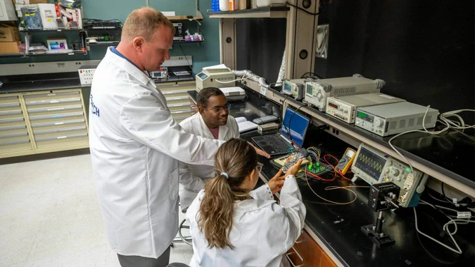 Three scientists in white coats gather around a counter and examine a handheld device attached to red and yellow cords.