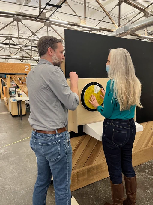 A woman and a man speak as they examine a and yellow and blue circular object affixed to a wooden board in a workshop.