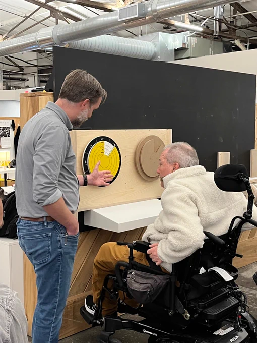 Two men, one standing and one in a wheelchair, examine two circular objects, one yellow and blue and the other brown, affixed to a wooden board.