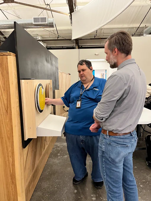 Two men stand near two circular objects, one yellow and blue and the other brown, affixed to a wooden board in a workshop. One of the men is touching the yellow and blue circular object.