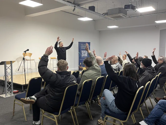 Speaker at Work Station networking event gesturing enthusiastically on stage in front of an audience raising their hands.