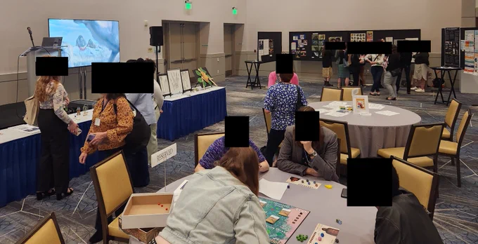 Busy conference hall with tables where people are playing a board game and art is displayed on long tables and on posters