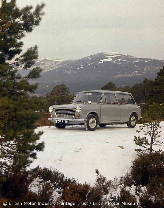 A colour photograph of a grey Austin 1100 Countryman in Scotland. The vehicle is situated in a three-quarter view facing facing the camera with trees and foliage around the bottom and sides of the photograph. In the background mountains can be seen. 