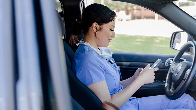 A healthcare professional in scrubs is sitting in a car, looking at a smartphone.