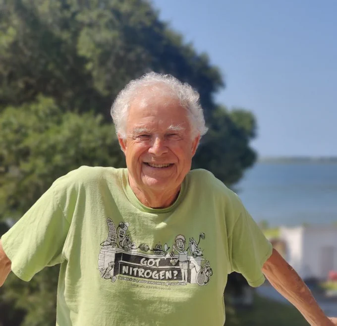 Photo of Dr. Hans Paerl standing in front of a blue sky, green trees, and blue water. He is smiling and wearing a green shirt that says "got nitrogen?"