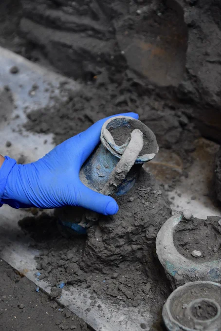 A conservator’s gloved hand gently removing the bronze jug from the marble top. A bronze lamp with moon-shaped decoration sits next to the jug