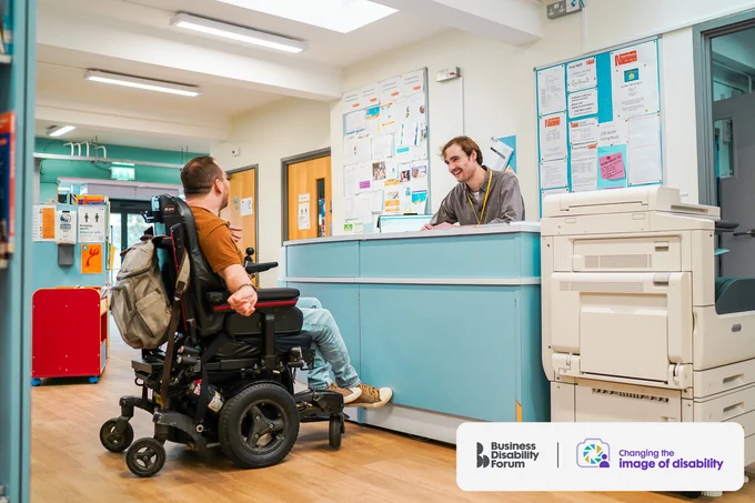 Person with cerebral palsy who uses a wheelchair talks to a receptionist in a library. The Business Disability Forum 'Changing the image of disability' logo is in the lower right-hand corner of the image.