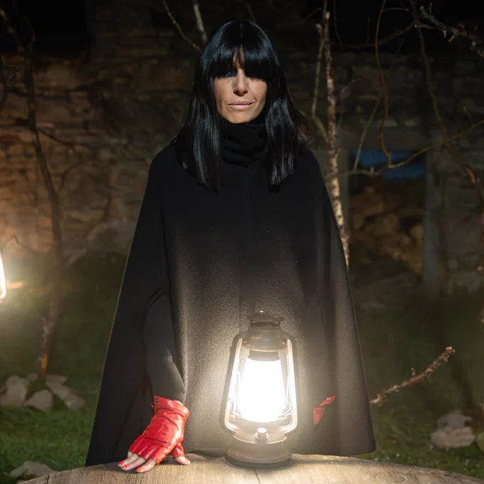 The Traitors host Claudia Winkleman stands outside at night. Wearing a black cloak and red, fingerless gloves, she rests her hand on a wooden table. Three outdoor lamps illuminate the scene with a golden glow. 