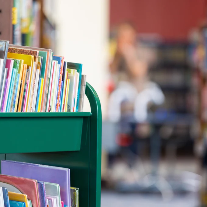 A trolley of books in a library in the foreground with people blurred out in the background.